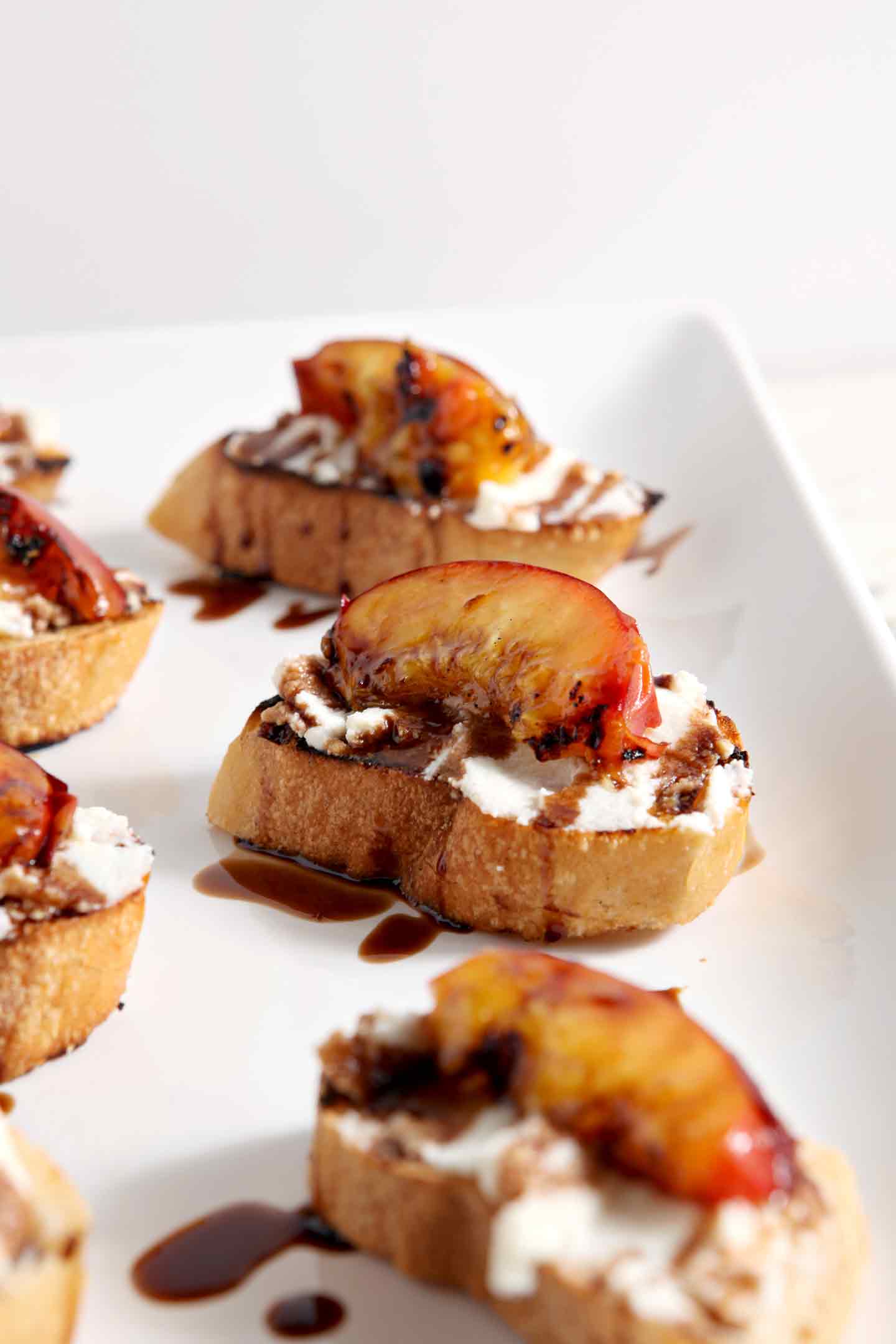 Grilled Peach Bruschetta sit on a white platter, drizzled with a balsamic reduction, before serving