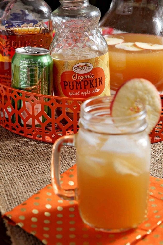 A drinking jar of an orange drink in front of a bottle of Sprouts Pumpkin Spiced Apple Cider, ginger ale, bourbon and a pitcher in an orange basket