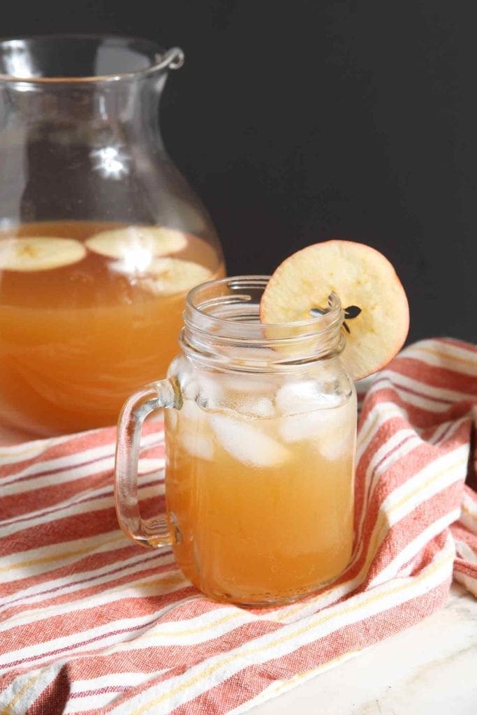 A jar of an orange drink garnished with an apple round sits on an orange-yellow-white striped kitchen towel with a pitcher of the drink behind it