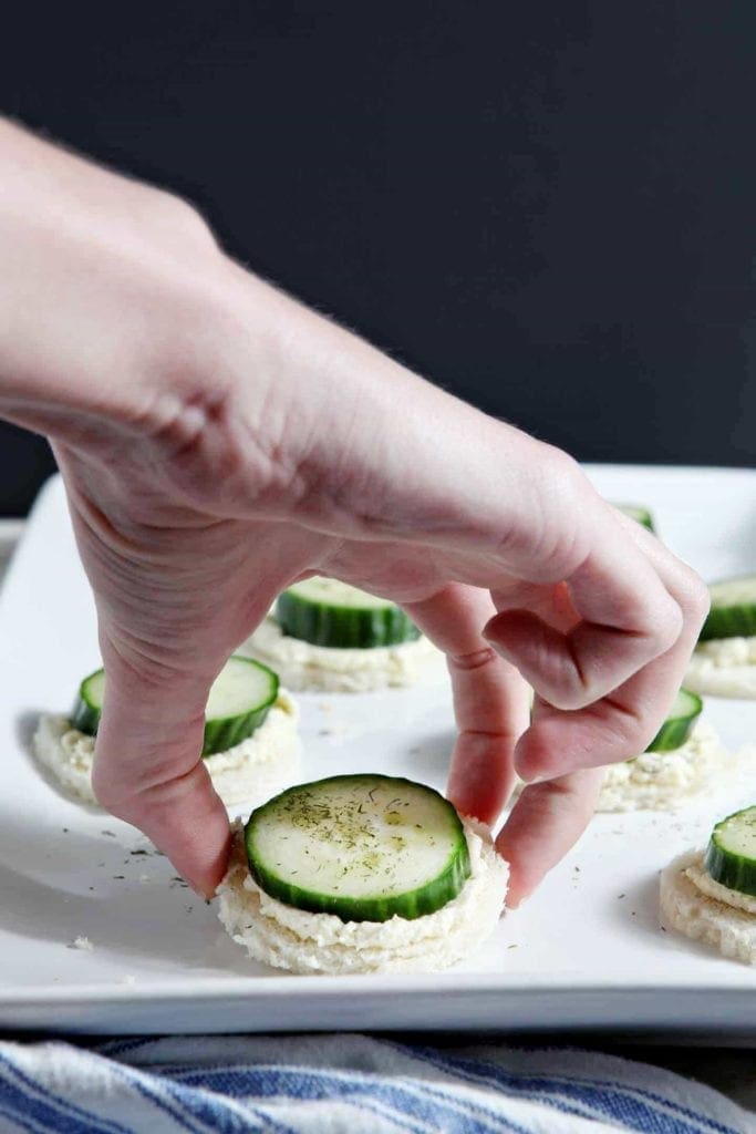 Hand picking up cucumber finger sandwich from white platter 