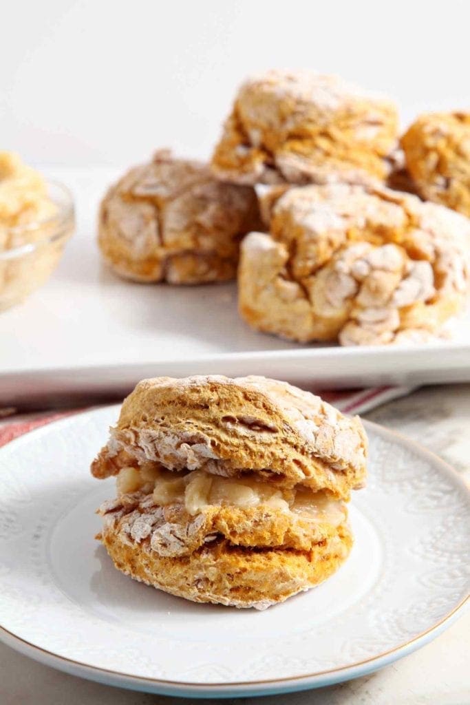 A Vegan Pumpkin Biscuit sits on a white plate in front of a platter of more biscuits