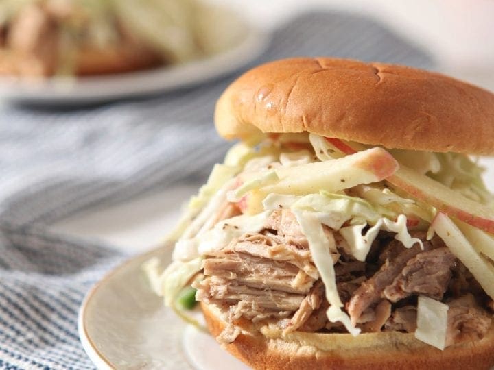 Close up of an Apple Cider Pulled Pork Sandwich with slaw on a white plate on top of a blue striped towel