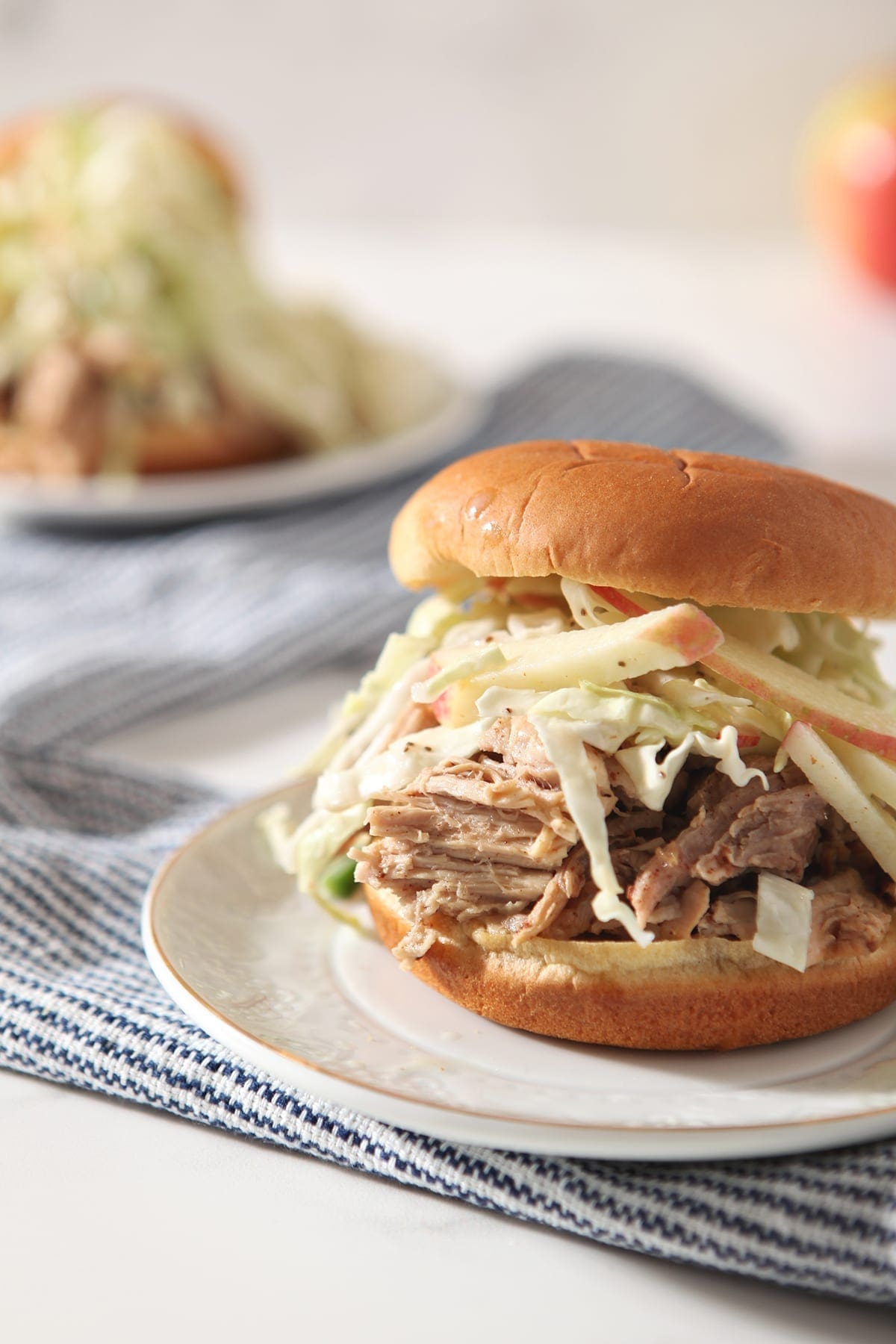 Close up of an Apple Cider Pulled Pork Sandwich with slaw on a white plate on top of a blue striped towel