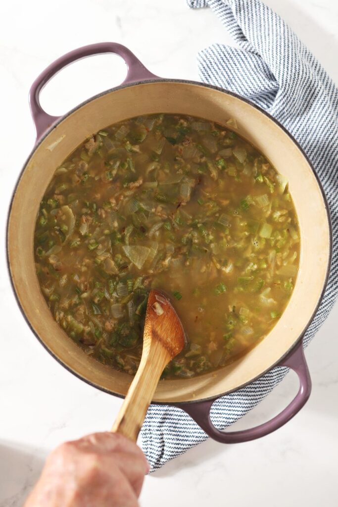 A wooden spoon stirs the Cajun trinity vegetables and stock in a Dutch oven