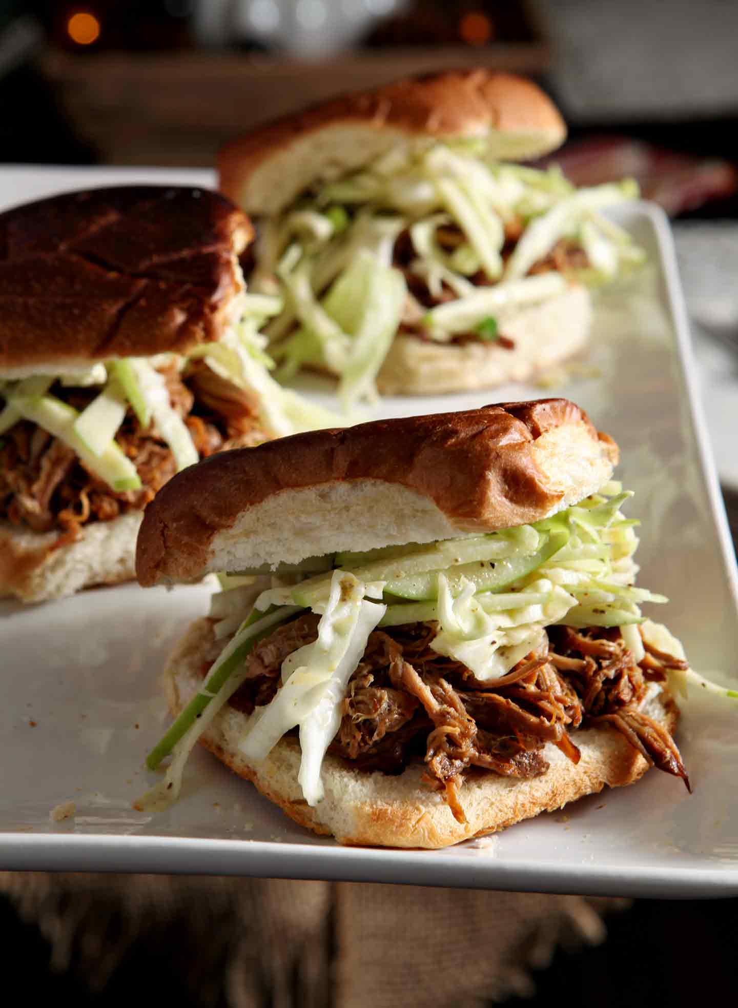 Three Apple Cider Pulled Pork with Apple Cabbage Slaw are served on buns on a white platter