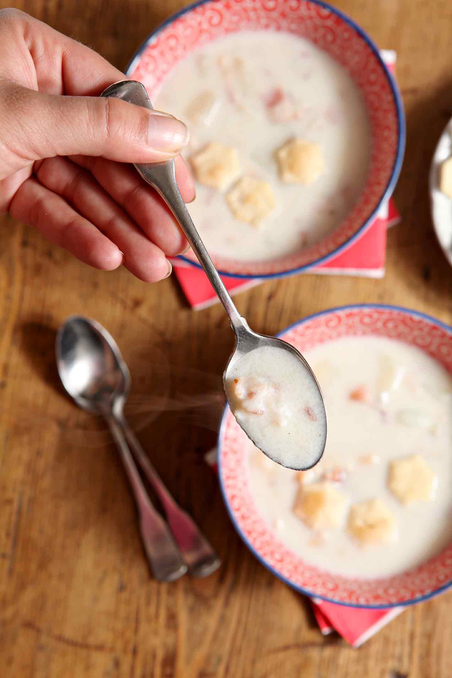 Hand holding a spoon over two bowls of Bacon Potato Clam Chowder 