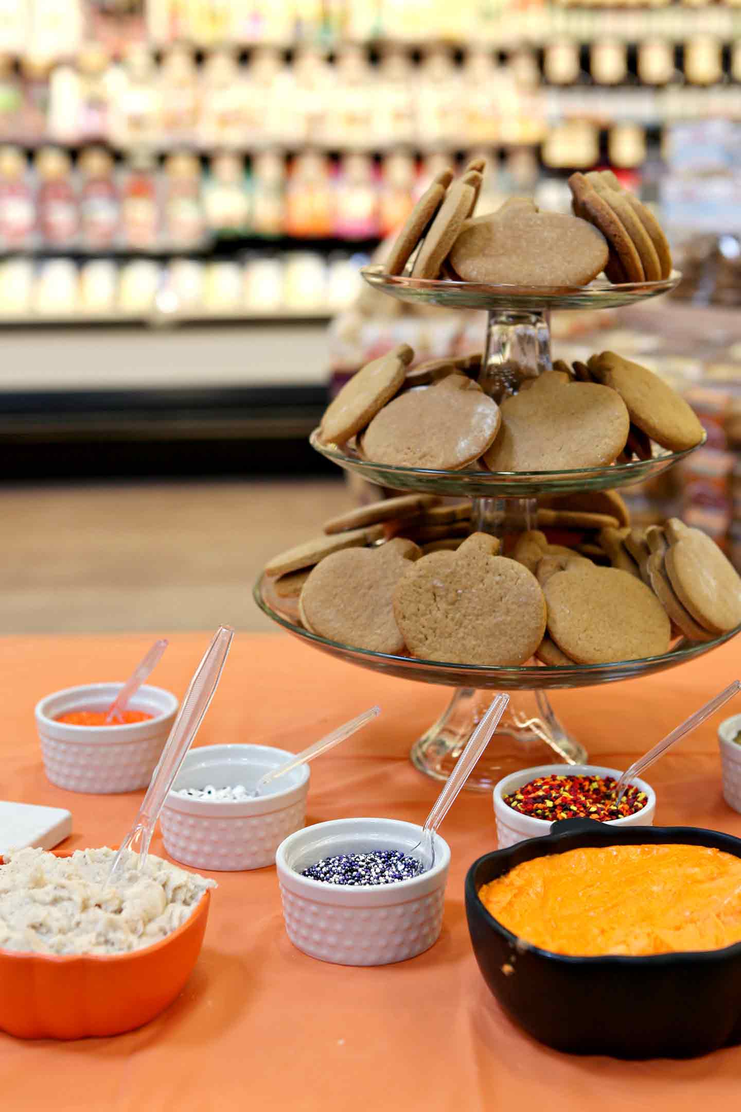 A cupcake stand holds undecorated pumpkin-shaped cookies and is surrounded by white bowls holding sprinkles and decorating items inside of a Sprouts