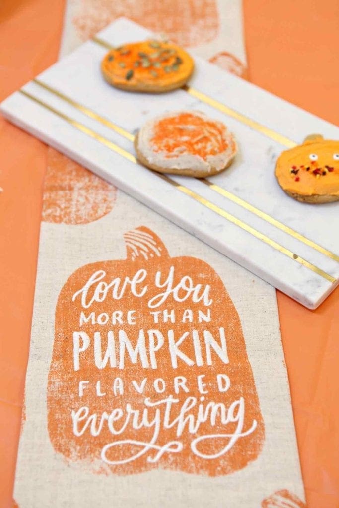 A towel stating 'love you more than pumpkin flavored everything' sits on an orange tablecloth next to decorated pumpkin cutout cookies at Sprouts