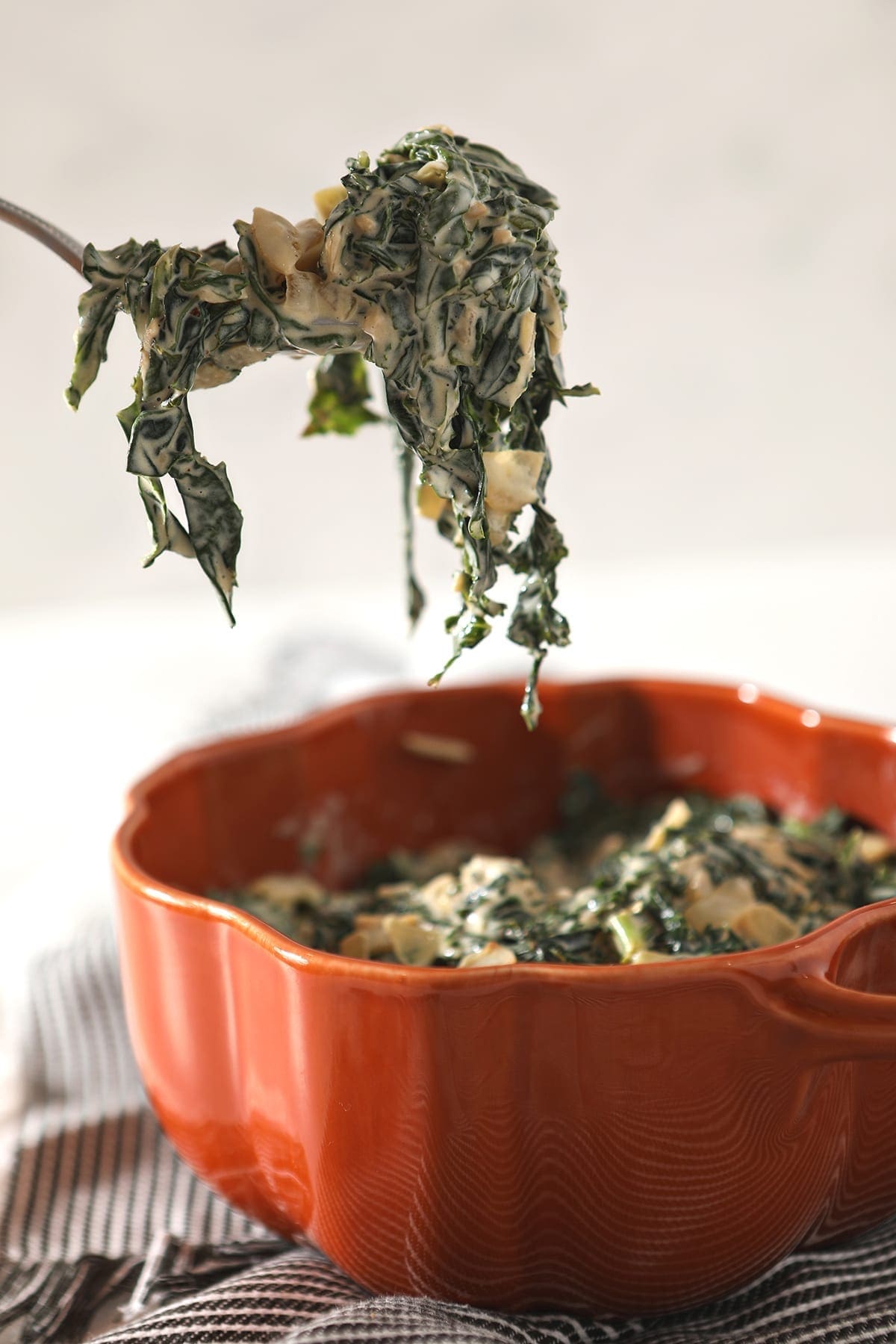 A spoon lifts a spoonful of Creamy Kale out of an orange serving dish that sits on a gray striped towel