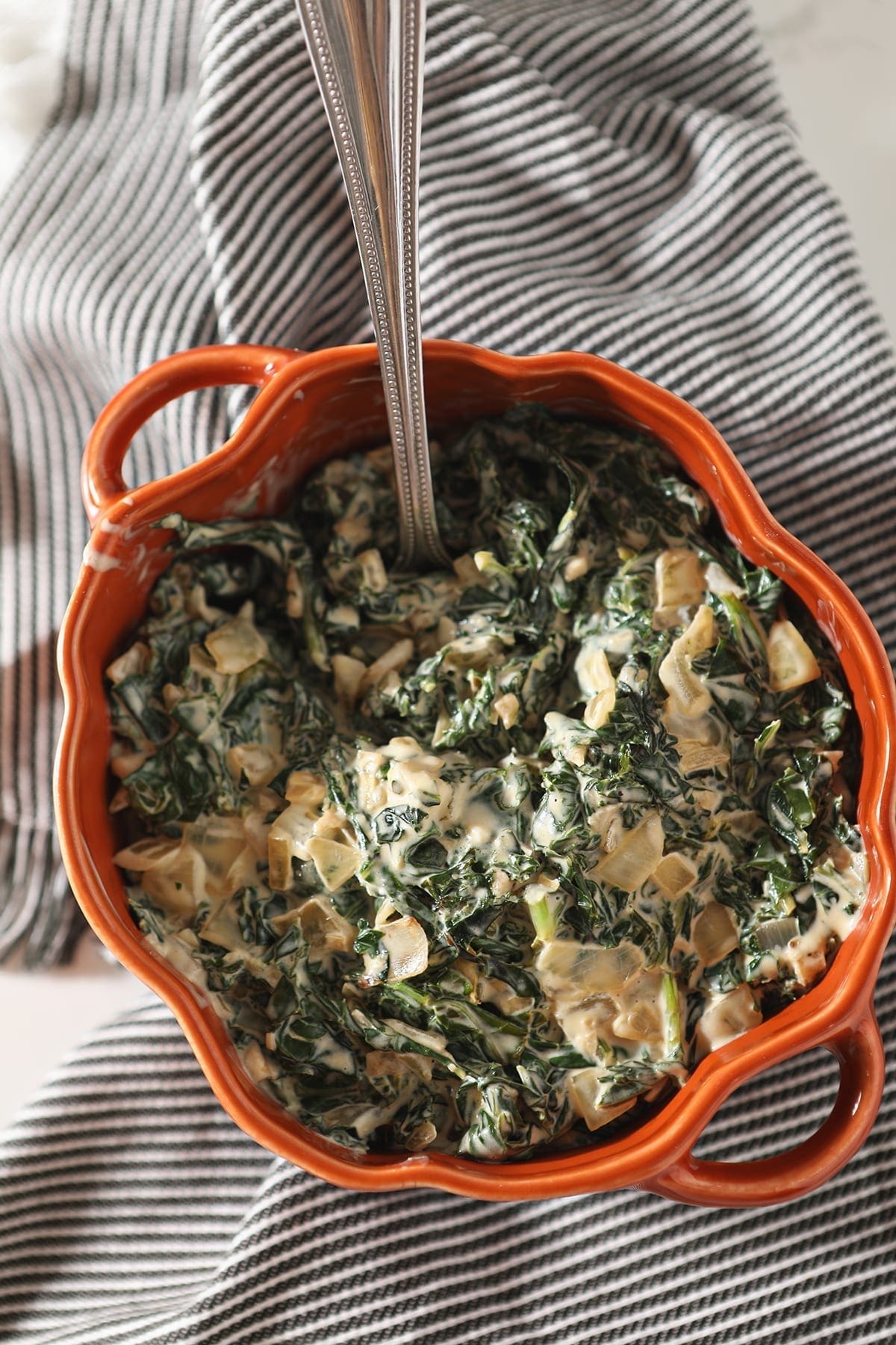 A silver spoon sits in an orange dish of Creamy Kale sitting on a gray striped towel