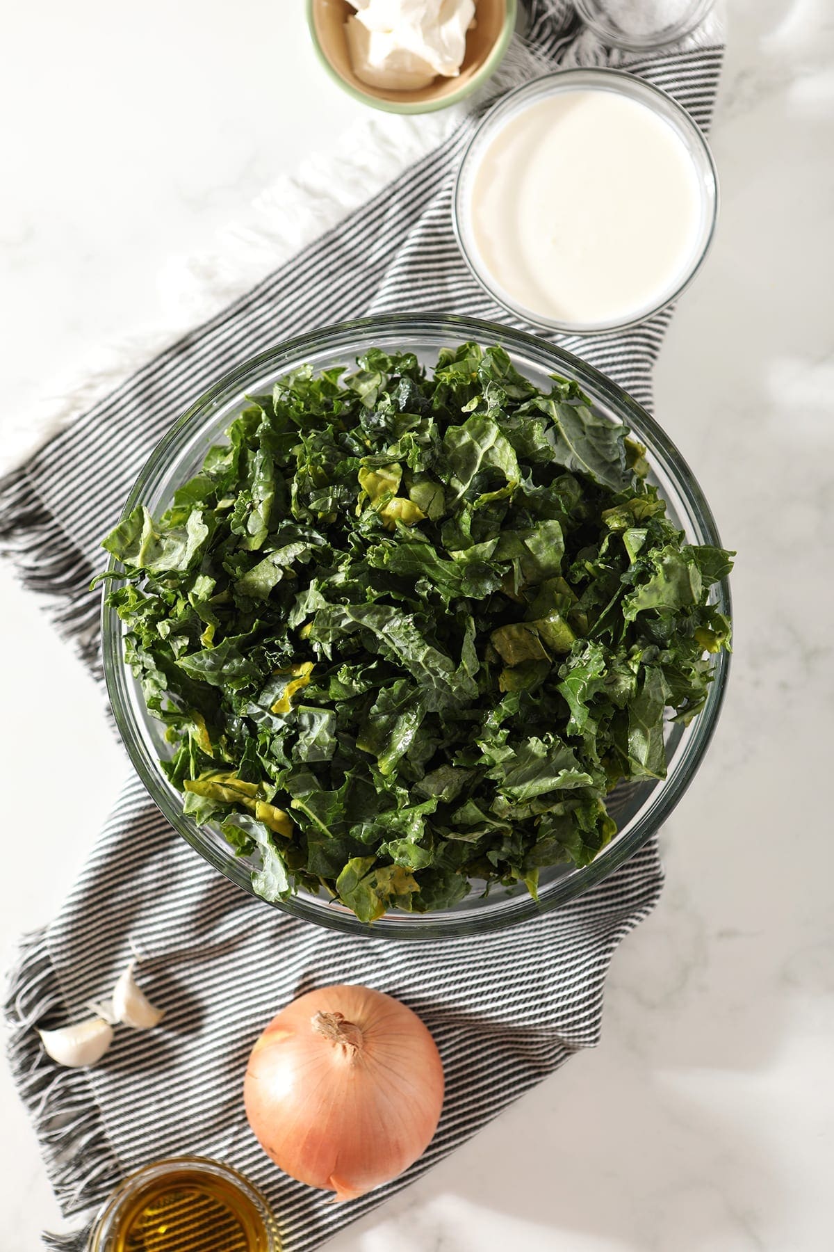 Chopped fresh kale in a bowl sitting on top of a gray striped towel, sitting next to an onion, garlic and other ingredients