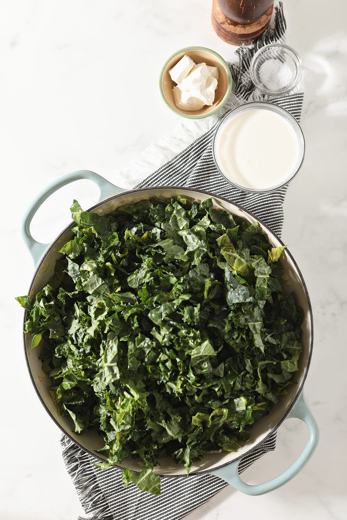 Chopped kale in a braiser pan on a gray striped towel next to cream cheese, cream, salt and pepper