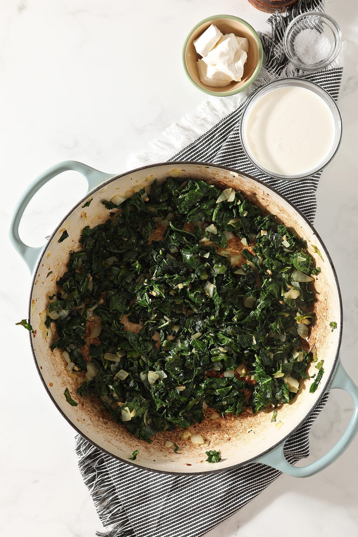 Overhead of sauteed kale and garlic in a pan sitting on a gray striped towel next to cream and cream cheese