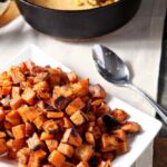 A white plate holds Bourbon Butter Sweet Potatoes with a skillet of cornbread in the background