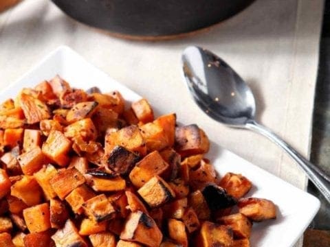 A white plate holds Bourbon Butter Sweet Potatoes with a skillet of cornbread in the background