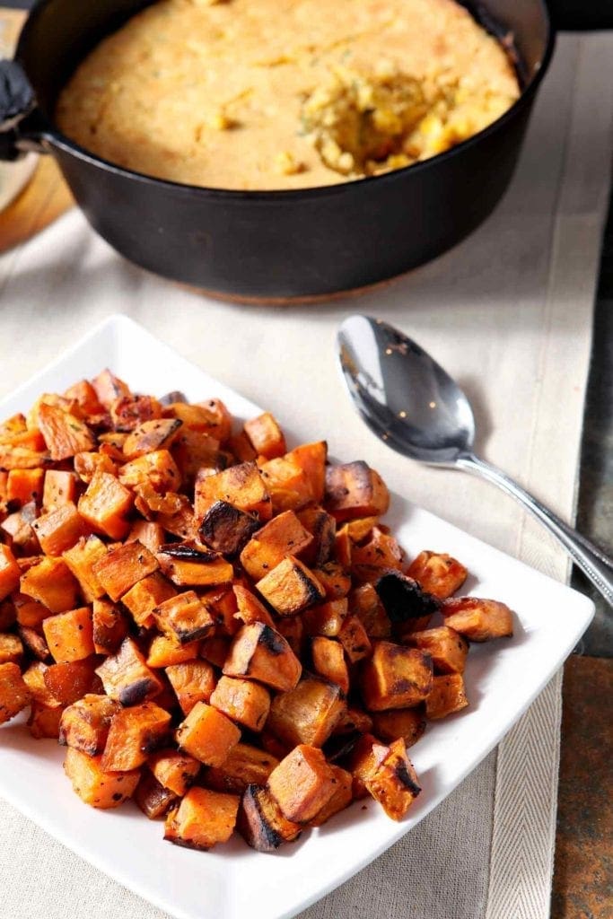 A white plate holds Bourbon Butter Sweet Potatoes with a skillet of cornbread in the background