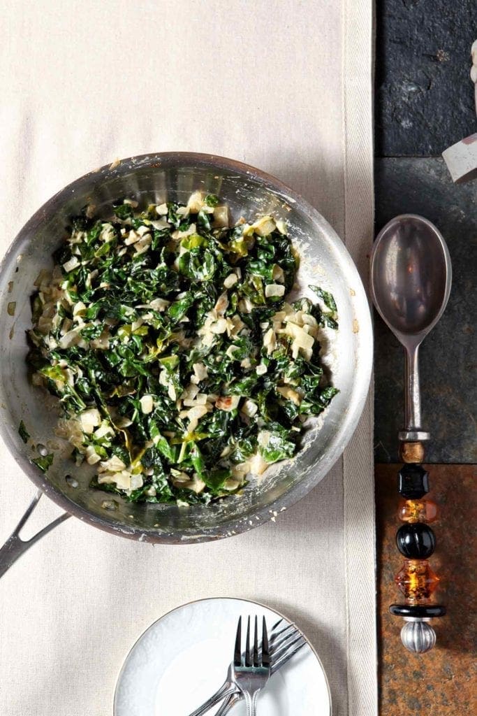 A silver skillet of a creamed kale recipe sits on a beige tablerunner next to a silver serving spoon and a white plate holding forks