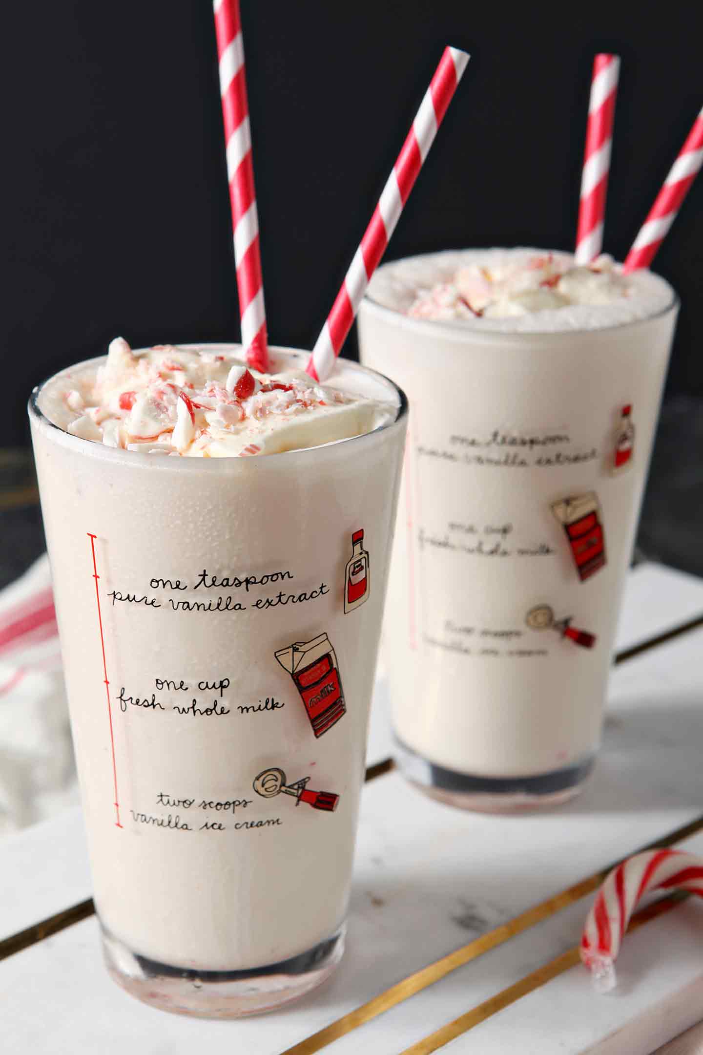 Two pint glasses hold peppermint milkshakes, garnished with extra candy cane crumbles and red striped straws