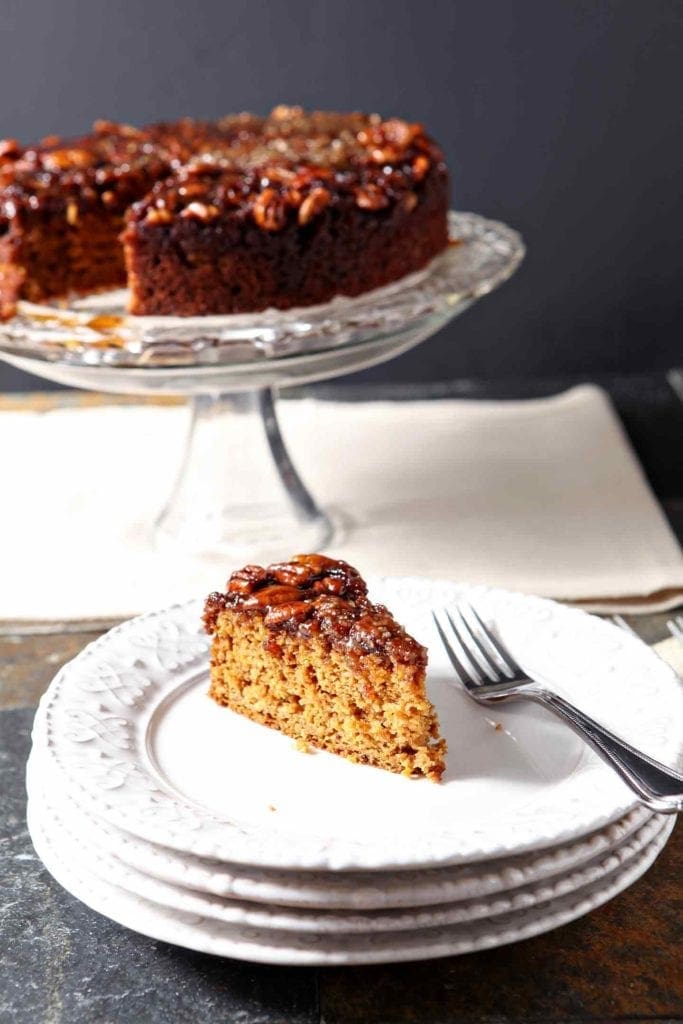 A slice of pumpkin cake with a praline topping sits on a white plate in front of a cake stand holding the rest of the dessert