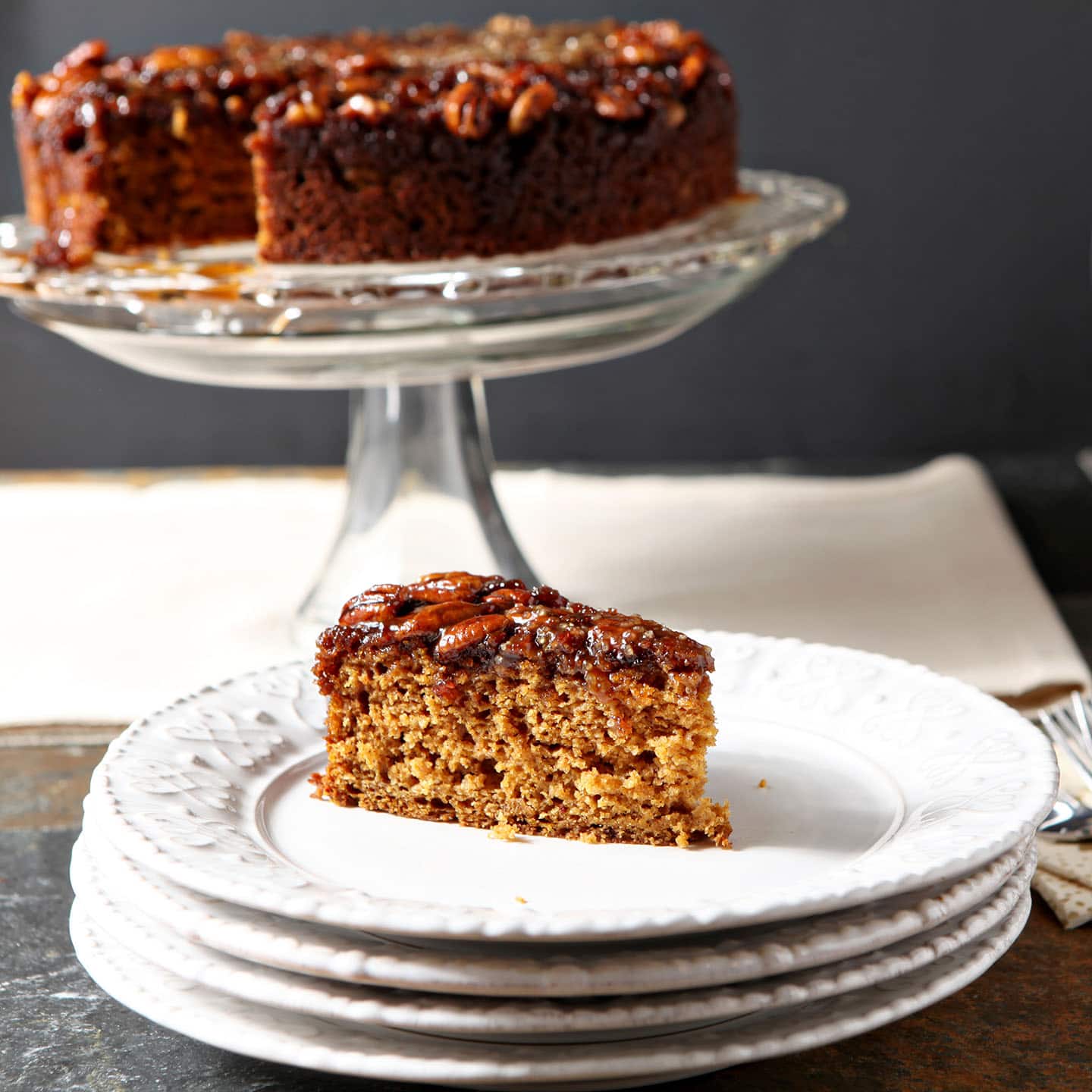 A slice of pumpkin cake with a praline topping sits on a white plate in front of a cake stand holding the rest of the dessert