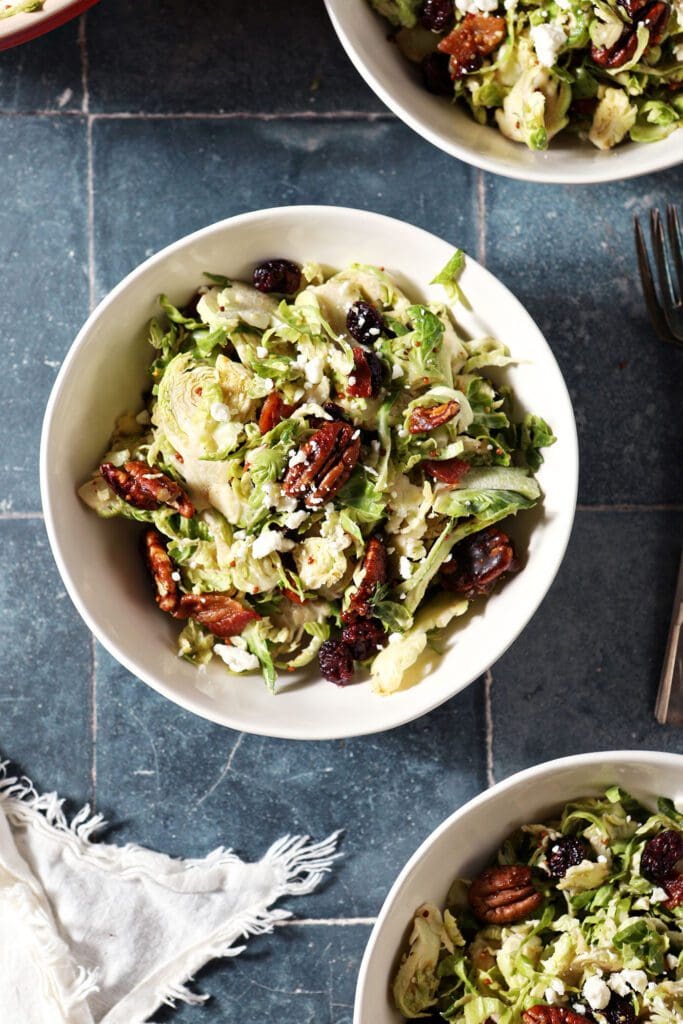 Three bowls holding individual servings of Cranberry Feta Brussels Sprout Salad on a dark blue tiled surface