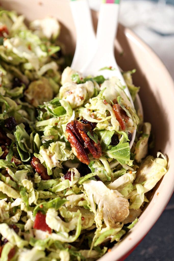 Close up of salad tongs in a bowl with a Cranberry Feta Brussels Sprout Salad with Candied Pecans and Warm Bacon Vinaigrette