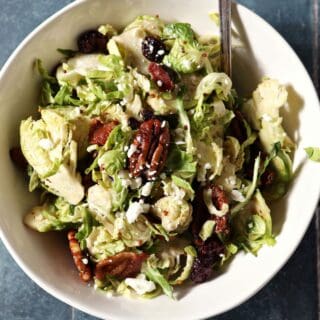 Close up of a bowl holding a serving of Cranberry Feta Brussels Sprout Salad with a fork on a dark blue tiled surface