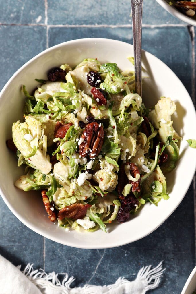 Close up of a bowl holding a serving of Cranberry Feta Brussels Sprout Salad with a fork on a dark blue tiled surface