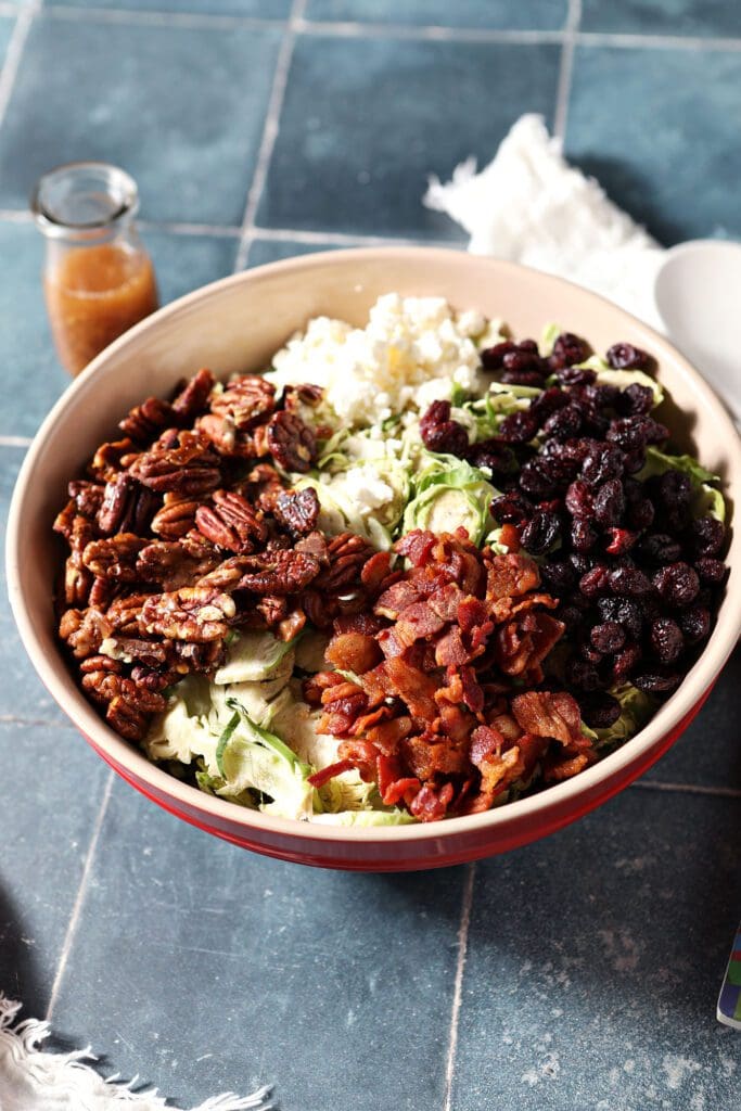 A large red mixing bowl holding individual ingredients to make a cranberry feta Brussels sprout salad on a blue tile surface next to a container of salad dressing