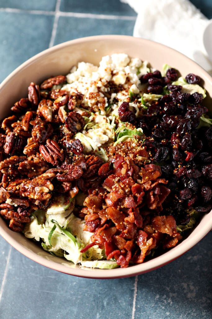 Cranberry Feta Brussels Sprout Salad with Candied Pecans and Warm Bacon Vinaigrette in a large red bowl on a blue tiled surface before tossing