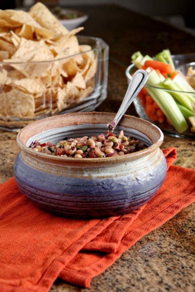 A bowl holding Black Eyed Pea Dip is served alongside other appetizers at a New Year's Day party