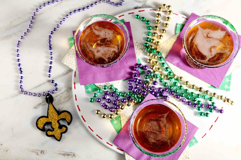 Overhead of three dessert martinis on purple napkins surrounded by purple, gold and green beads on marble