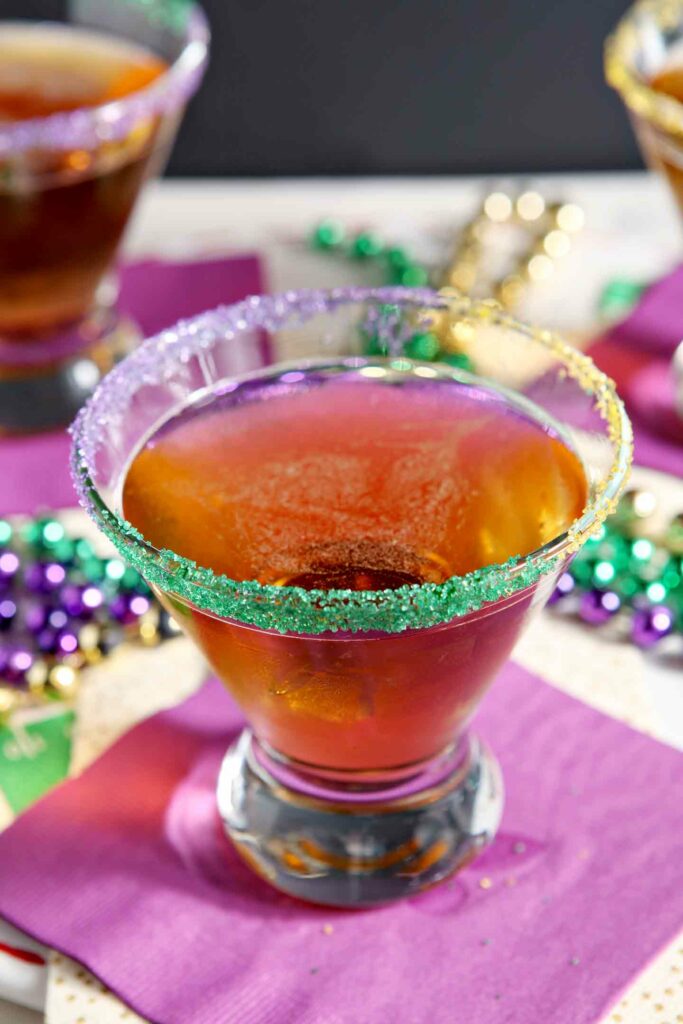 Close up of a king cake martini on a purple napkin, surrounded by festive mardi gras beads