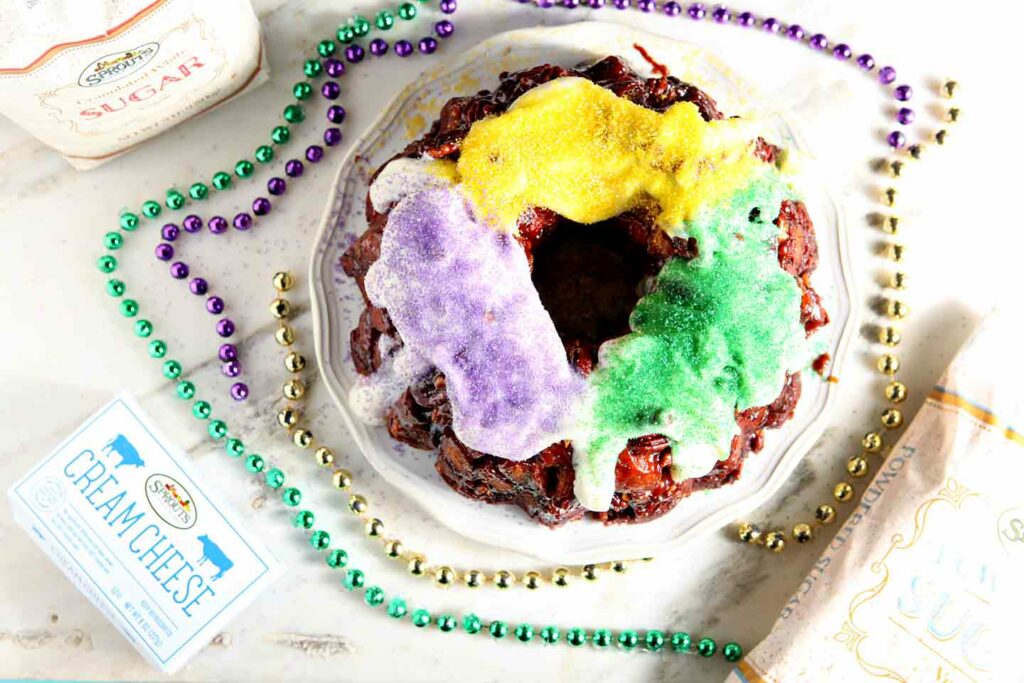 Overhead image of a cinnamon roll king cake on a marble slab next to cream cheese, powdered sugar and beads