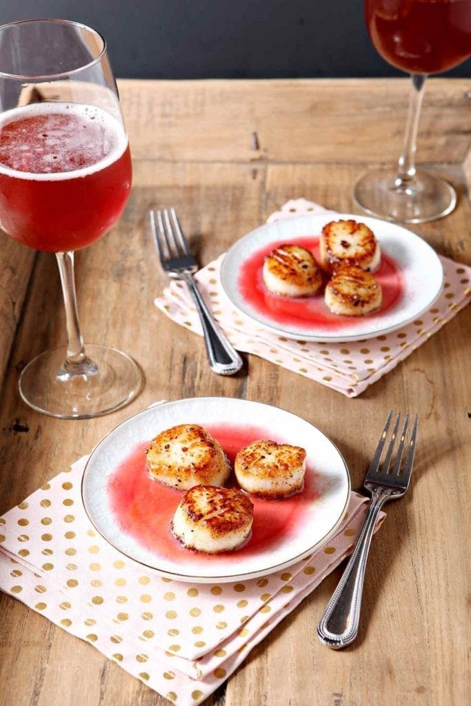 Two plates holding scallops with a red gastrique sit on a wooden tray next to a red drink