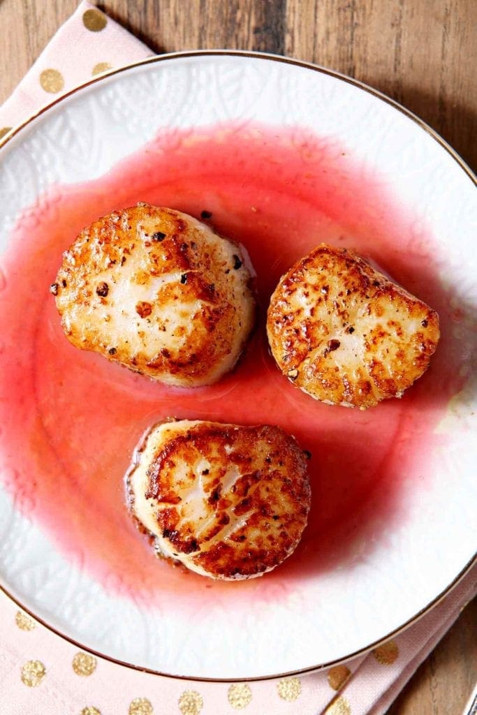 Three seared scallops sit on a white plate with raspberry gastrique on a wooden surface