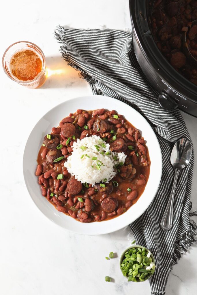 A bowl of Slow Cooker Red Beans and Rice sits next to a glass of beer and a slow cooker