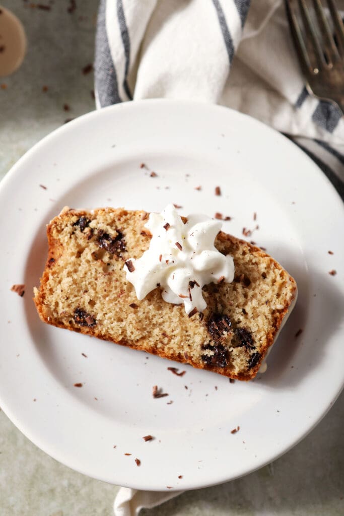 A slice of chocolate chip loaf cake topped with whipped cream and chocolate shavings on a plate