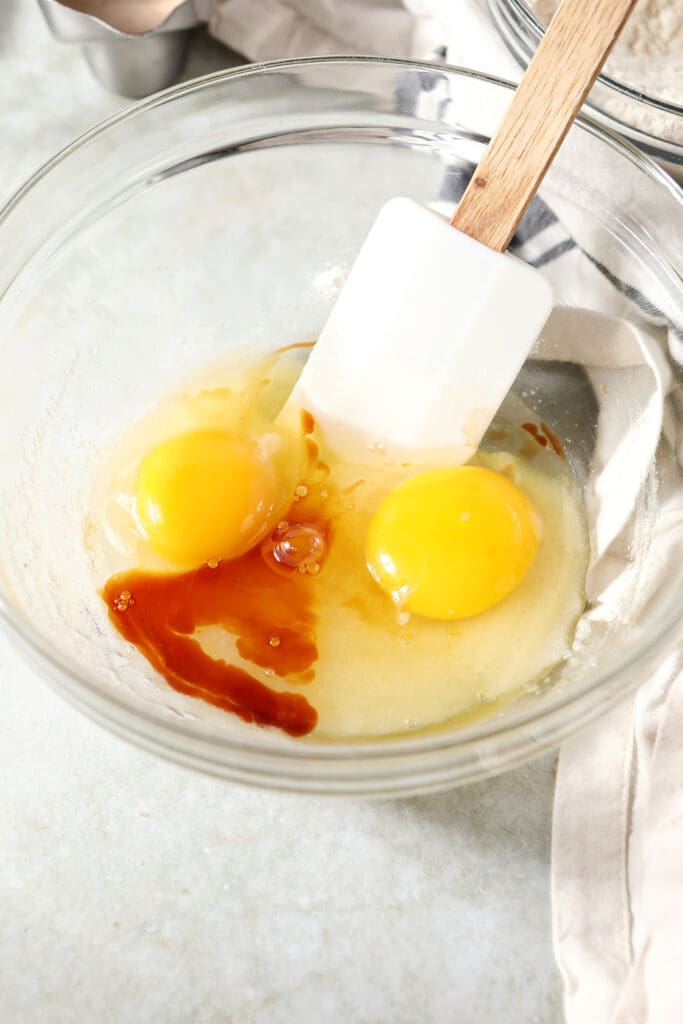 Eggs and vanilla added to granulated sugar in a bowl with a rubber spatula