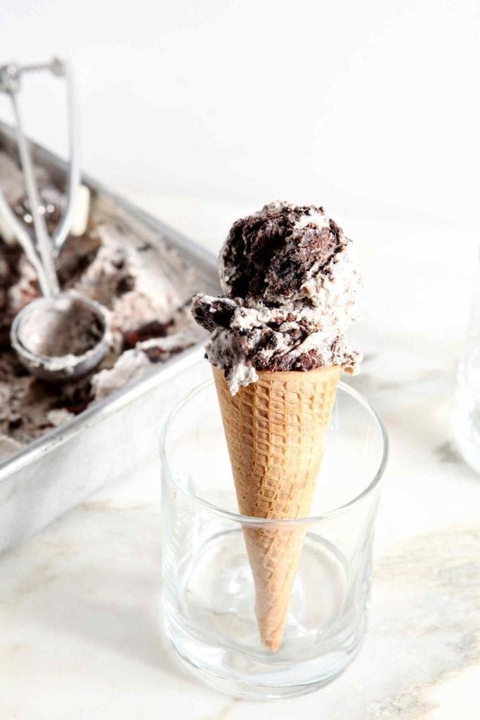 A scoop of No Churn Fudge Brownie Ice Cream sits atop a sugar cone in a glass with the carton of ice cream behind it.