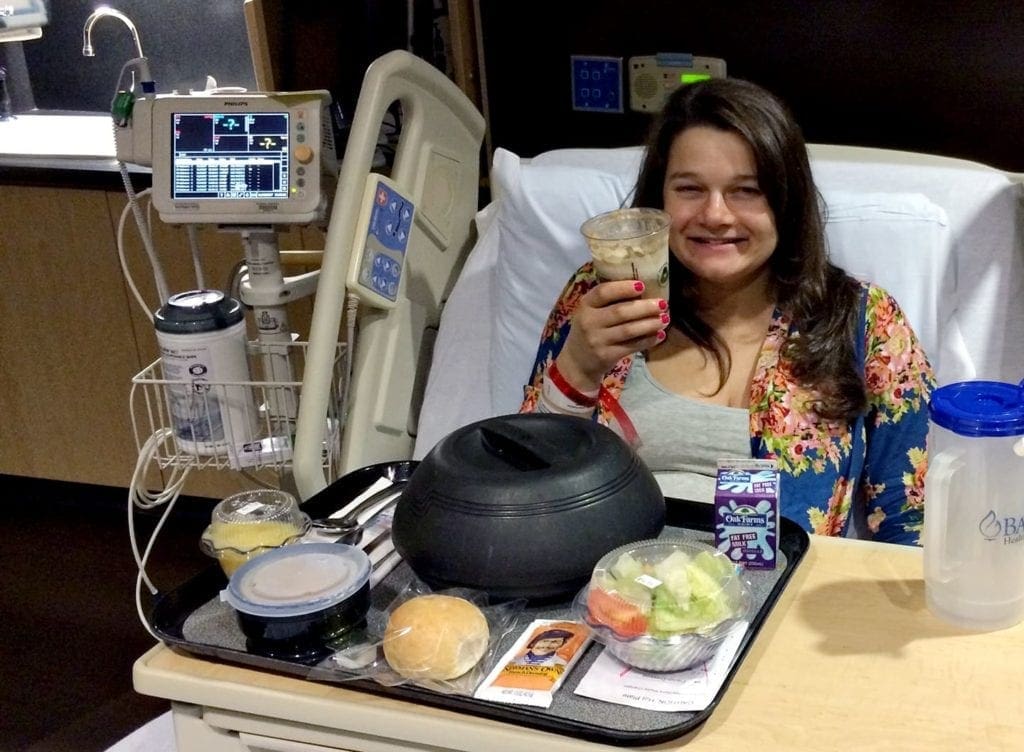 A smiling woman sits in a hospital bed holding a root beer float with a tray of food in front of her