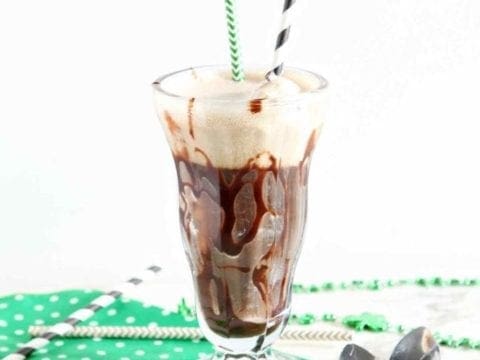 A single parfait glass holds a beer float on top of a green and white polka dot cloth next to straws and bistro spoons