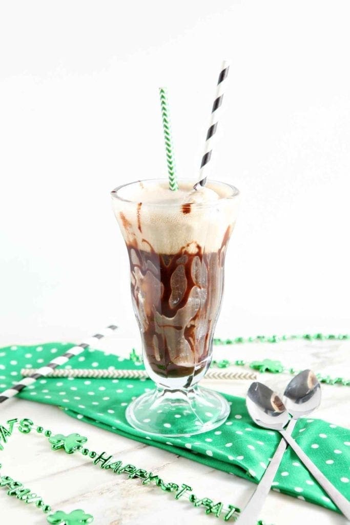 A single parfait glass holds a beer float on top of a green and white polka dot cloth next to straws and bistro spoons