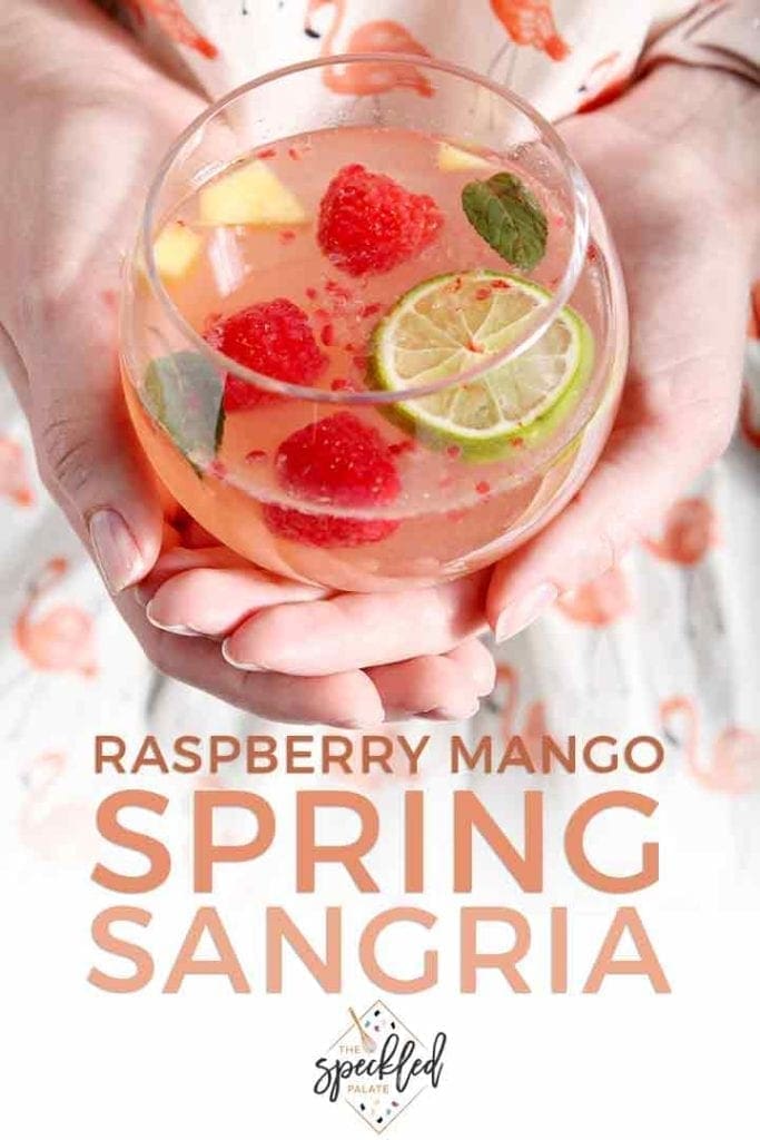 A woman in a flamingo shirt holds a glass of mango raspberry white wine sangria with the text 'raspberry mango spring sangria'