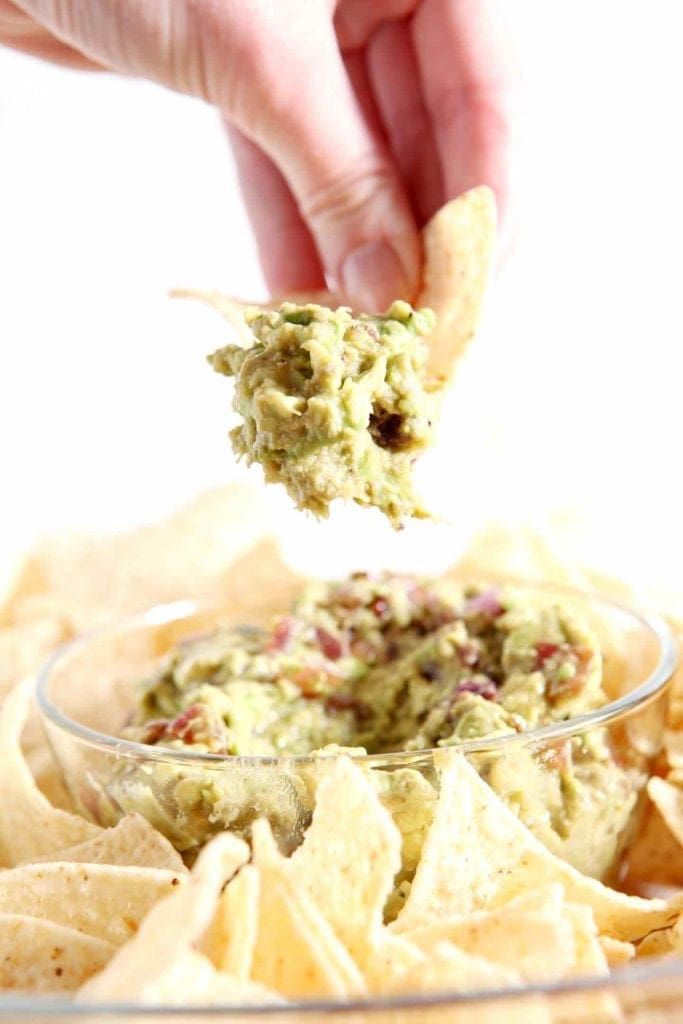 Caramelized Onion Guacamole being scooped out of a serving platter with a white corn tortilla chip before eating