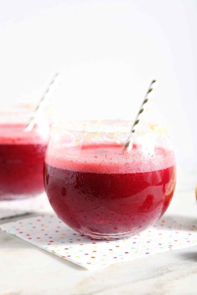 Two ruby red Cherry Lemonade Margaritas, served with gold striped straws on confetti napkins, are served on a white marble platter.