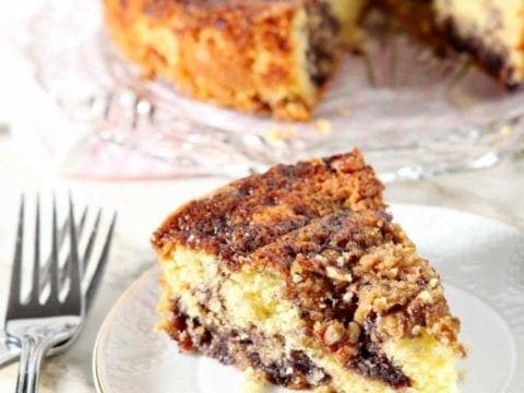 A slice of Lemon Blueberry Coffee Cake sits on a small white plate with the entire cake in the background, along with forks.
