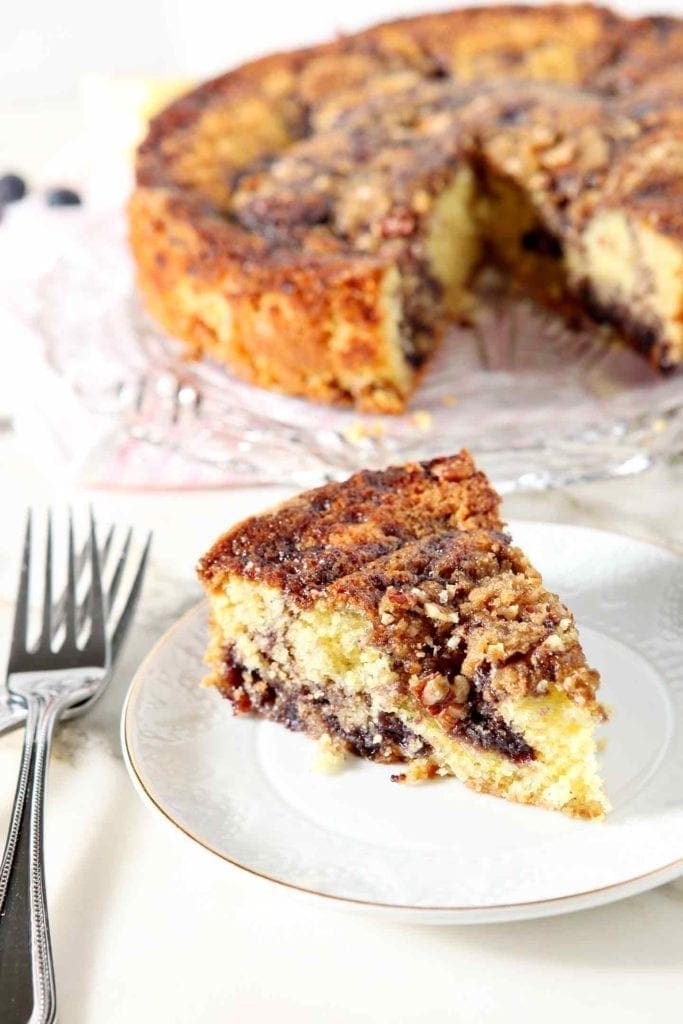 A slice of Lemon Blueberry Coffee Cake sits on a small white plate with the entire cake in the background, along with forks.