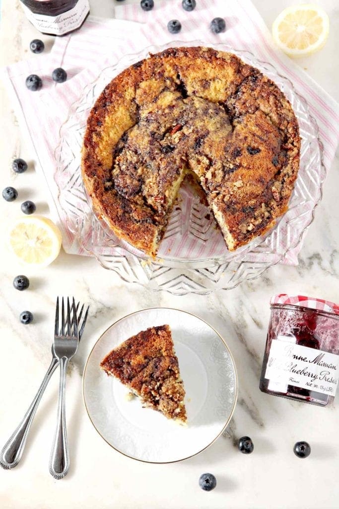 A slice is served on a plate next to a coffee cake and a jar of blueberry preserves