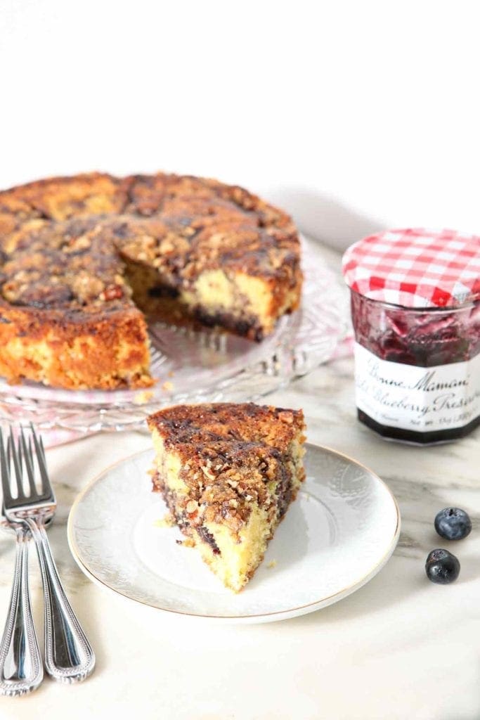 A slice of Lemon Blueberry Coffee Cake is served on a white plate, with the rest of the cake in the background